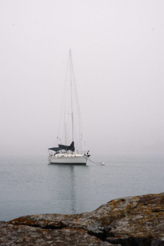 A white sailboat with a black canopy is anchored on calm water in foggy weather, with a rocky shoreline visible in the foreground. by Justin Salem Meyer