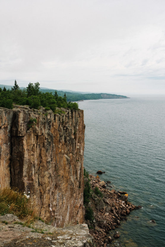 A steep, rocky cliff covered with green trees overlooks a calm, expansive body of water under an overcast sky. The shoreline and distant hills are visible in the background. by Justin Salem Meyer