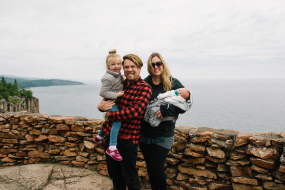 A smiling family of four poses by a stone wall overlooking a large body of water. One adult holds a young child, while the other holds a baby wrapped in a blanket. It appears to be a cool, cloudy day. by Justin Salem Meyer