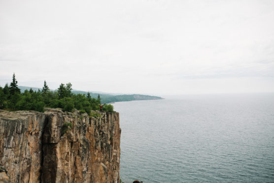 A person stands on the edge of a tall, rocky cliff overlooking a large, calm body of water, with trees and vegetation along the cliff and distant shoreline under a cloudy sky. by Justin Salem Meyer