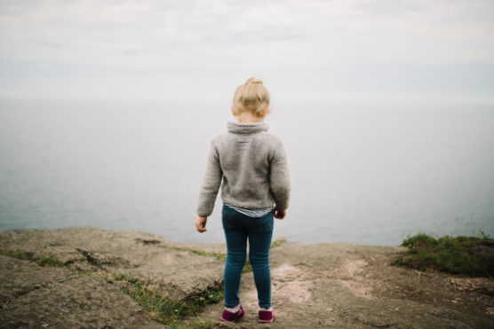 A young child in a gray sweater and jeans stands on rocky ground facing a calm, foggy body of water, with their back to the camera under a cloudy sky. by Justin Salem Meyer