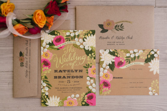 A floral-themed wedding invitation suite with beige, green, pink, and white flower designs, including an invitation, RSVP card, and envelopes. A flower crown with orange and pink blooms rests above the cards on a wooden surface. by Justin Salem Meyer