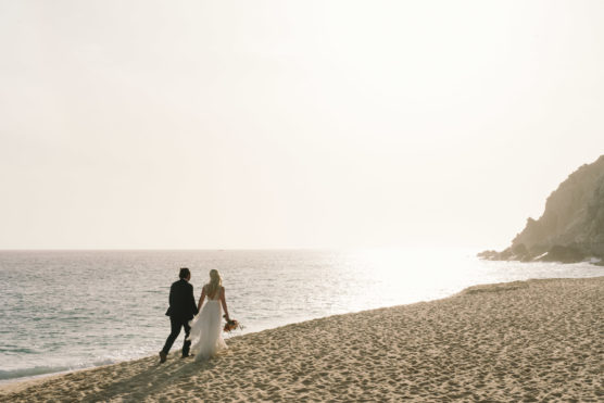A bride and groom walk hand-in-hand along a sandy beach at sunset, with gentle waves and a rocky cliff in the background under a bright, hazy sky. by Justin Salem Meyer