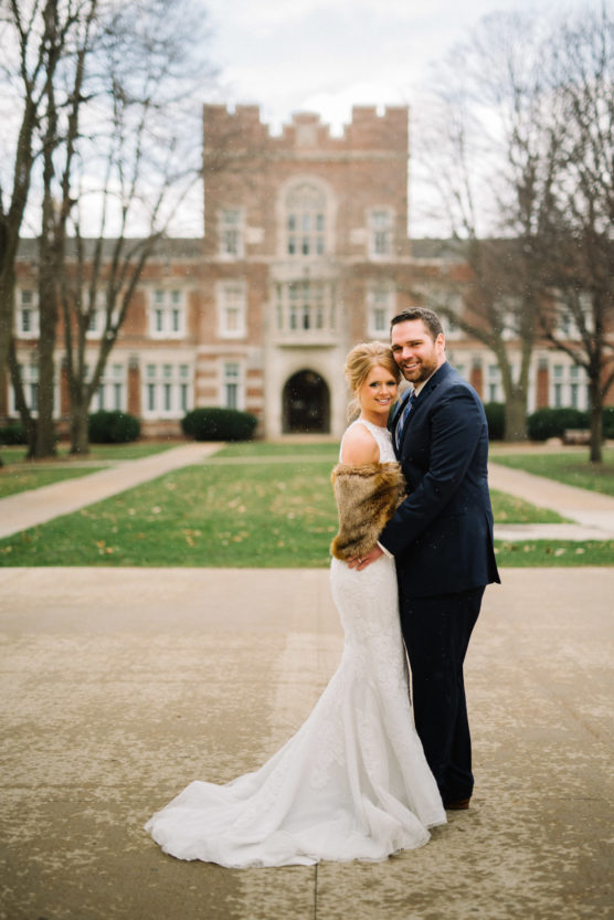 A bride in a white gown and fur shawl stands with a groom in a navy suit, smiling and embracing each other outdoors in front of a large brick building on a cloudy day. by Justin Salem Meyer