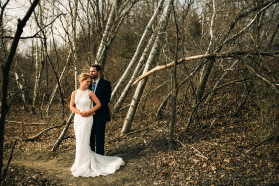 A bride and groom stand embracing on a forest path surrounded by bare trees and fallen leaves. The bride wears a white lace gown, and the groom is in a dark suit, both looking thoughtfully into the distance. by Justin Salem Meyer
