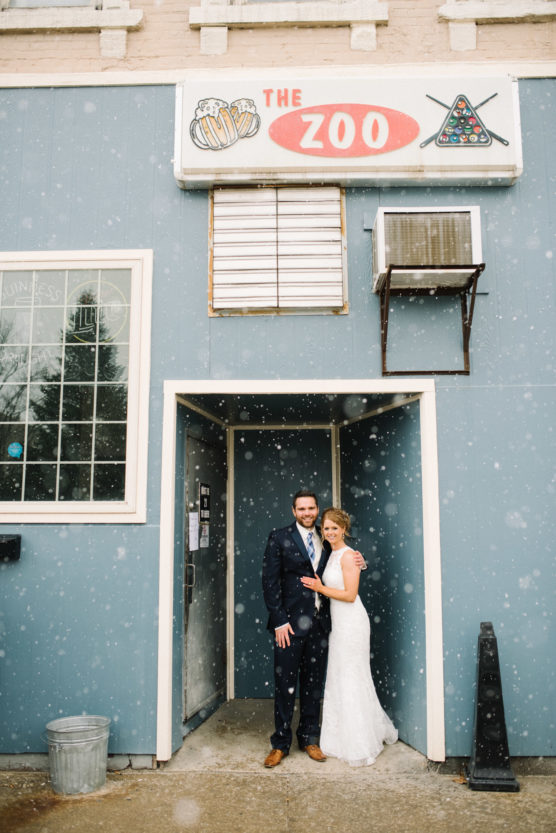 A bride and groom stand smiling together in the doorway of a blue building with a sign reading The Zoo above them, as snow falls around them. The bride wears a white gown; the groom is in a dark suit. by Justin Salem Meyer