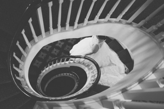 A black and white photo of a bride in a flowing wedding dress seen from above, standing on a carpeted landing in the middle of a spiral staircase with white railings. by Justin Salem Meyer