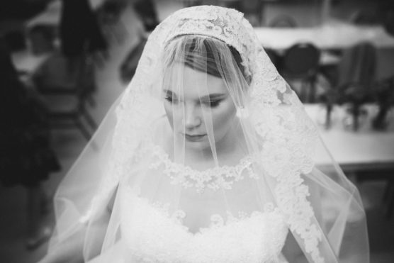 A black and white photo of a bride wearing a lace wedding dress and a matching lace veil, looking down with a serene expression. Tables and chairs are blurred in the background. by Justin Salem Meyer