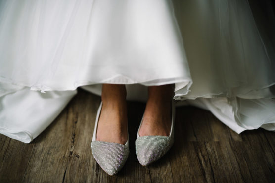 A bride wearing a white wedding dress stands on a wooden floor, with only her lower legs and sparkling, rhinestone-covered shoes visible beneath the hem of her gown. by Justin Salem Meyer