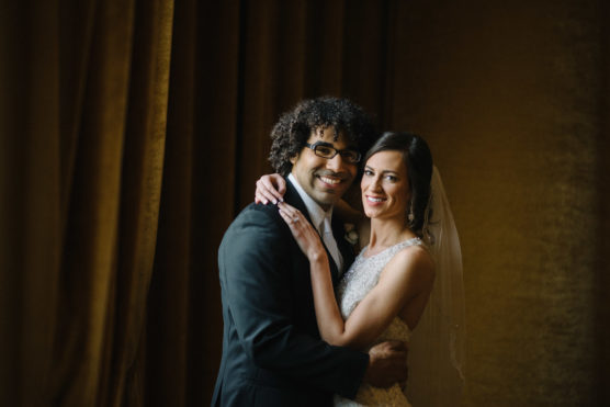 A bride and groom smile and embrace in front of dark brown curtains. The bride wears a white wedding dress and veil, while the groom wears a black suit and glasses. Warm light highlights their faces. by Justin Salem Meyer