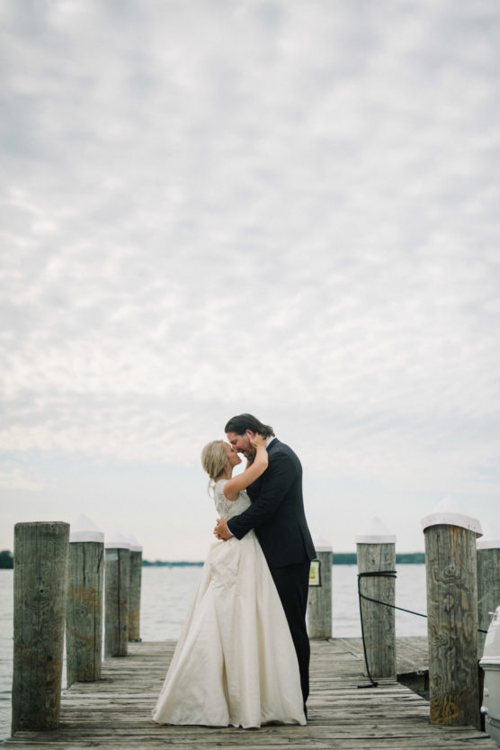 A bride and groom embrace and kiss on a wooden dock over a calm lake, with a cloudy sky above and water in the background. by Justin Salem Meyer