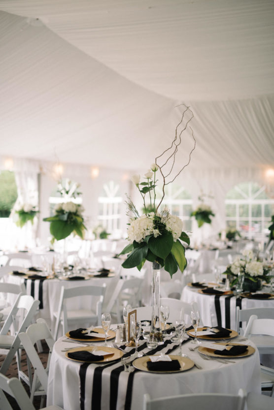 Elegant wedding reception setup in a white tent, featuring round tables with black and white striped runners, gold-accented plates, and tall floral centerpieces with white flowers and greenery. White chairs are neatly arranged. by Justin Salem Meyer