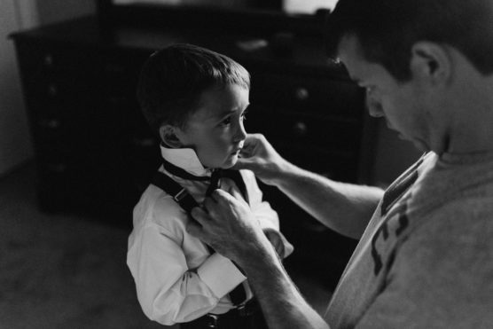 A man helps a young boy adjust his tie and collar. The boy, dressed in a white shirt and suspenders, looks forward seriously as the man gently fixes his outfit in a softly lit room. by Justin Salem Meyer