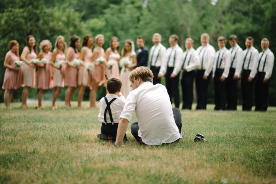 A man and a young boy sit on grass facing a wedding party, with bridesmaids in pink dresses and groomsmen in white shirts and suspenders standing in a row outdoors. by Justin Salem Meyer