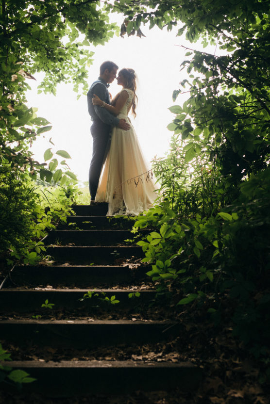 A couple in wedding attire stands embracing at the top of outdoor stone steps, surrounded by lush green foliage and framed by arching tree branches, with bright light in the background. by Justin Salem Meyer