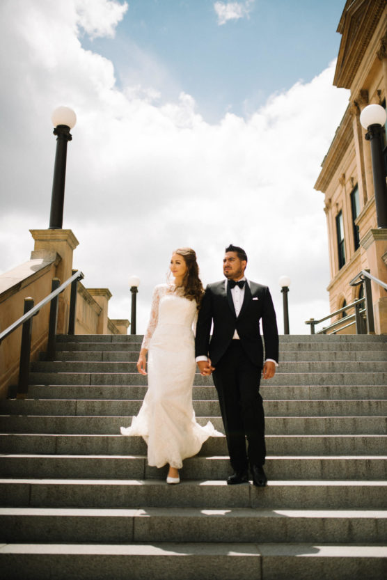 A bride in a long white lace gown and a groom in a black tuxedo walk down stone steps outside, holding hands, with lamp posts and a building in the background under a partly cloudy sky. by Justin Salem Meyer