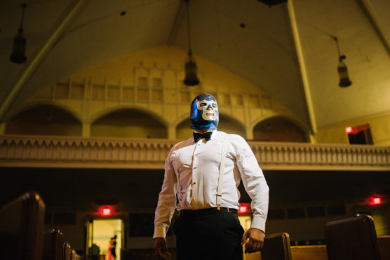 A person wearing a blue luchador wrestling mask, white dress shirt, and suspenders stands confidently in the aisle of a dimly lit hall or church, with high ceilings and hanging lights above. by Justin Salem Meyer