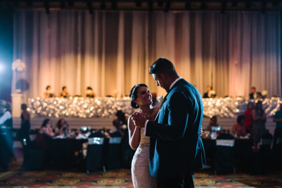 A bride and groom share their first dance at a wedding reception, smiling at each other on a dimly lit dance floor with guests seated in the background and string lights behind them. by Justin Salem Meyer