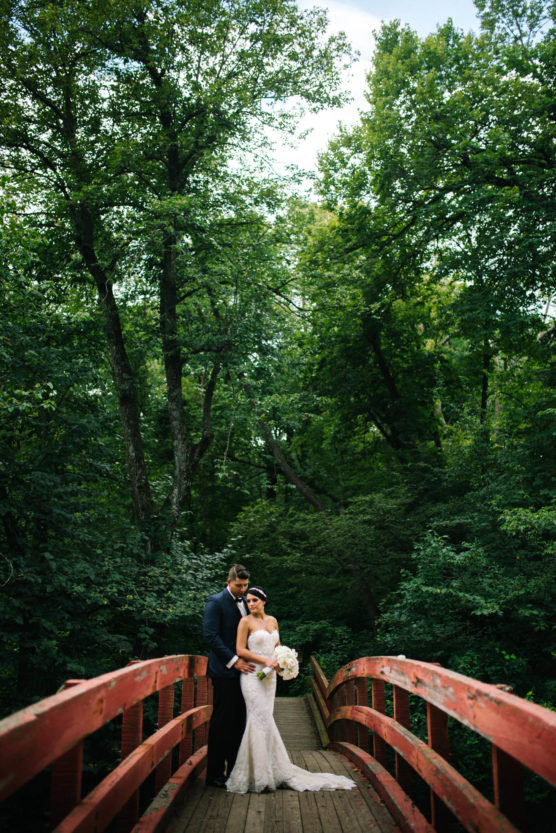 A bride and groom stand together on a rustic red wooden bridge surrounded by lush green trees, with the bride holding a bouquet and the groom embracing her from behind. by Justin Salem Meyer