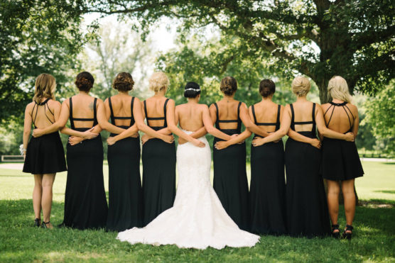 Eight women stand outdoors with their backs to the camera, arms linked. Seven wear matching black dresses, and one in the center wears a white wedding gown. They stand on grass beneath leafy trees in sunlight. by Justin Salem Meyer