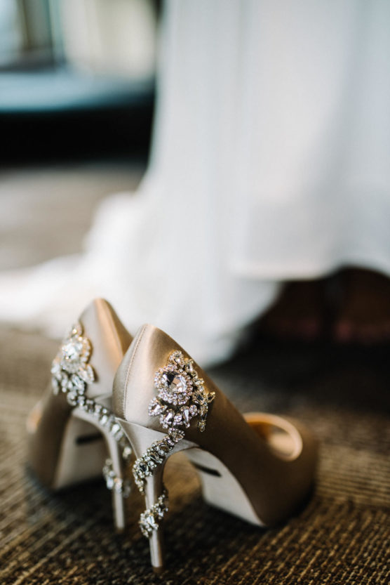 A pair of elegant high-heeled shoes adorned with sparkling rhinestone decorations, placed on a carpeted floor. A white dress is softly blurred in the background. by Justin Salem Meyer