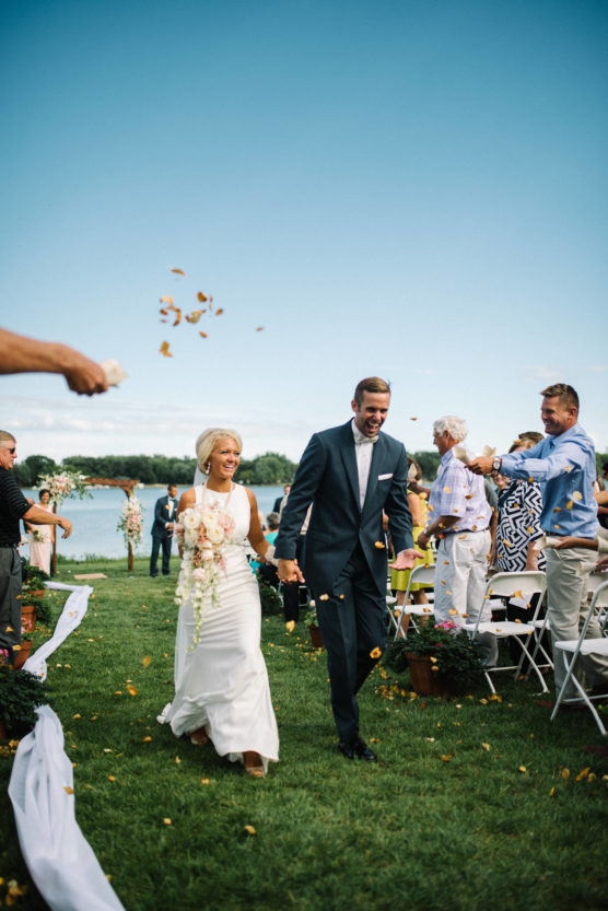 A bride and groom walk hand in hand outdoors as wedding guests toss flower petals. The couple is smiling, with rows of chairs and people on either side, and a lake and blue sky in the background. by Justin Salem Meyer