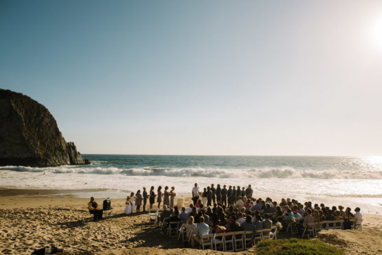 A wedding ceremony is taking place on a sandy beach with the ocean and waves in the background. Guests are seated in rows, and the wedding party stands in a semicircle facing the officiant. The sun shines brightly in the clear sky. by Justin Salem Meyer