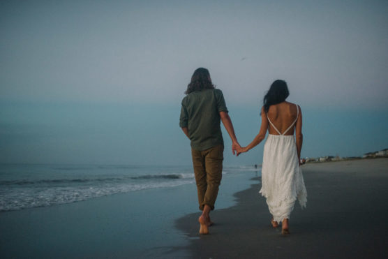 A couple walks barefoot on the beach at dusk, holding hands. The woman wears a white dress, and the man wears a green shirt and brown pants. The ocean is calm, and the sky is a soft blue-gray. by Justin Salem Meyer