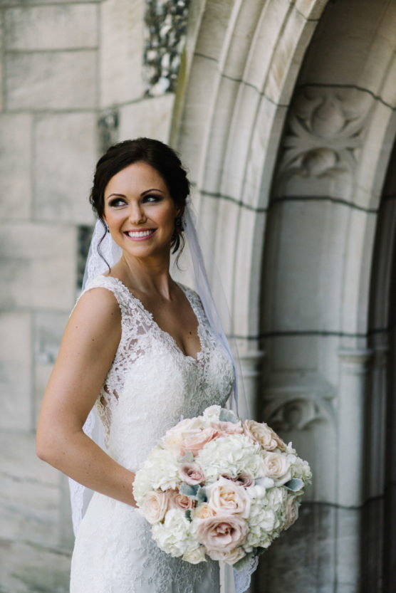 A bride in a lace wedding dress holds a bouquet of pale roses and hydrangeas, smiling and looking to the side, standing in front of a stone archway. by Justin Salem Meyer