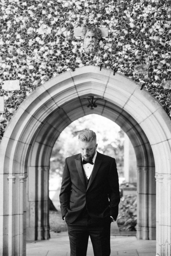 A man in a tuxedo stands under a stone archway with his hands in his pockets, looking down. The background features textured stonework and foliage seen through the arch. by Justin Salem Meyer