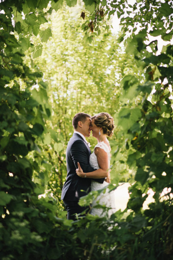 A bride and groom embrace surrounded by lush green foliage, standing close together and touching foreheads in a natural outdoor setting. by Justin Salem Meyer
