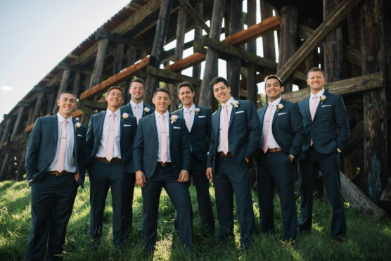 Nine men in matching blue suits and light pink ties stand smiling together outdoors on grass, with a large wooden bridge structure behind them, under a bright sky. by Justin Salem Meyer