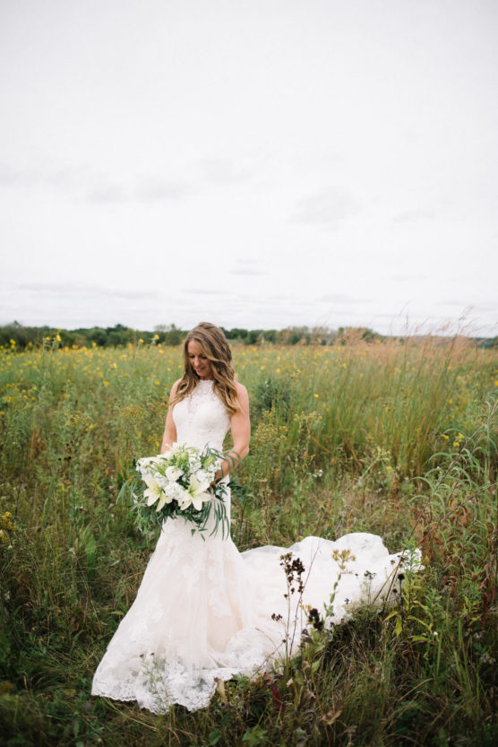 A bride in a white lace gown stands in a field of tall grass and wildflowers, holding a large bouquet of white flowers and greenery, looking down and smiling softly. by Justin Salem Meyer