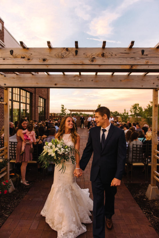 A bride in a white gown and a groom in a navy suit hold hands and smile at each other as they walk down an outdoor aisle under a wooden arbor, with guests seated on either side at sunset. by Justin Salem Meyer