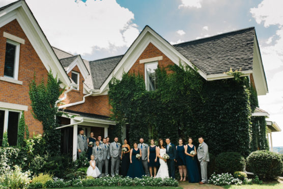 A large wedding party stands together outside a red brick house covered in ivy. The group includes people in grey suits and dark teal dresses, with the bride in white at the center, all posing for a photo on a sunny day. by Justin Salem Meyer