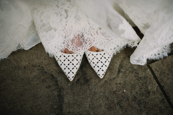 A close-up view of a bride’s white lace wedding dress partially covering her pointed white shoes with a geometric cut-out pattern, standing on a stone surface. by Justin Salem Meyer