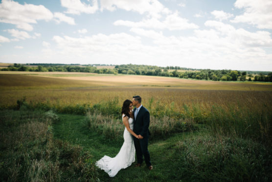 A bride and groom stand close together in a grassy field under a partly cloudy sky, with rolling farmland and green trees stretching out in the background. by Justin Salem Meyer