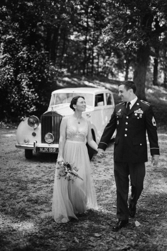 A bride in a flowing dress and a groom in a military uniform walk hand in hand, smiling at each other, with a vintage car parked behind them in a sunlit outdoor setting. by Justin Salem Meyer