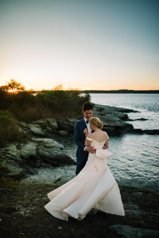 A bride and groom embrace on a rocky shoreline at sunset, with the bride’s white dress flowing and the ocean in the background. The sky glows with warm light, creating a romantic atmosphere. by Justin Salem Meyer