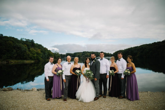 A wedding party stands on gravel by a calm lake, with the bride and groom in the center, surrounded by bridesmaids in purple dresses and groomsmen in white shirts and gray pants. Trees and cloudy sky are reflected in the water. by Justin Salem Meyer