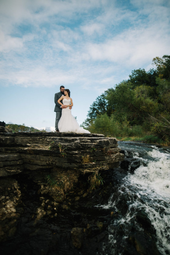 A bride and groom stand on a rocky ledge near a flowing river, surrounded by greenery and under a blue sky with scattered clouds. The bride wears a white dress; the groom is in a gray suit. by Justin Salem Meyer