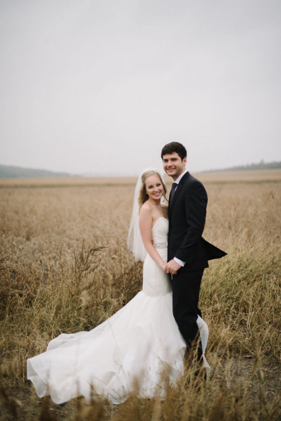 A bride in a white gown and veil and a groom in a black suit stand smiling together in a field of tall, golden grass under a cloudy sky. by Justin Salem Meyer