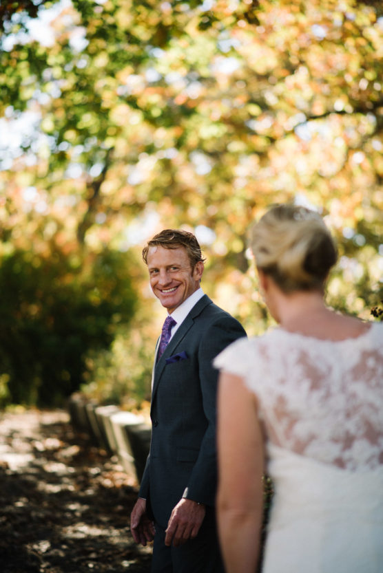A man in a suit smiles warmly at a woman in a white dress with lace details, outdoors with trees in the background showing autumn colors. Sunlight filters through the leaves, creating a soft, romantic atmosphere. by Justin Salem Meyer