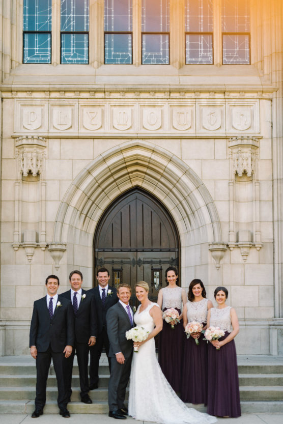 A wedding party of four men in suits and four women in dresses pose, smiling, in front of a large arched church doorway with Plymouth carved above it. The bride and groom stand at the center. by Justin Salem Meyer