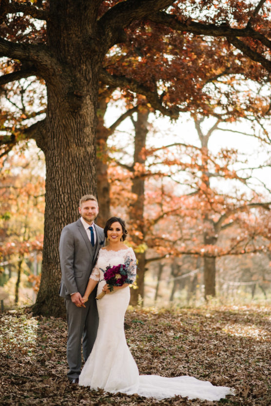 A bride in a white gown holding a bouquet and a groom in a gray suit stand smiling under a large tree with autumn leaves on the ground and colorful fall foliage in the background. by Justin Salem Meyer