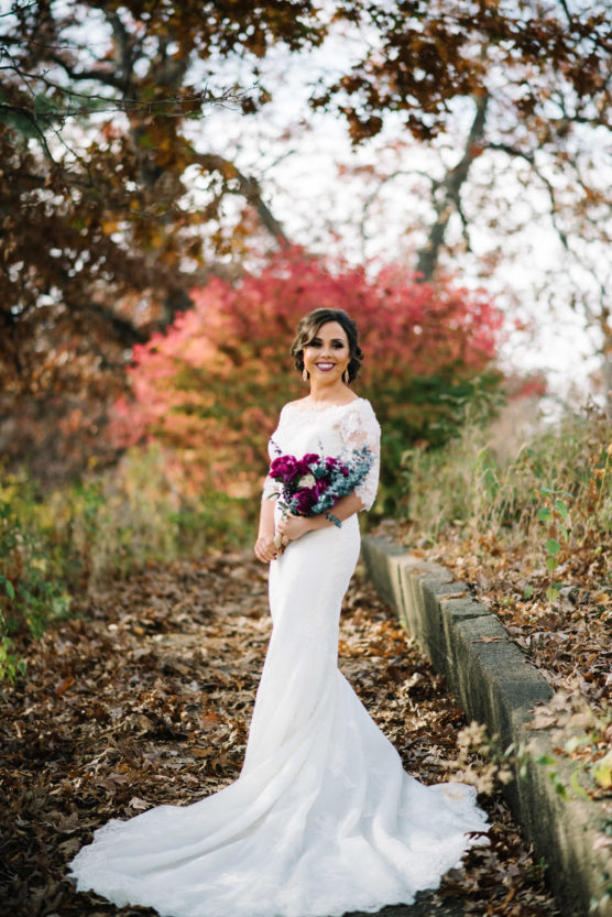 A bride in a white lace gown stands outdoors on a leaf-covered path, holding a bouquet of purple flowers. Autumn trees with red, orange, and brown leaves surround her, creating a vibrant, natural backdrop. by Justin Salem Meyer