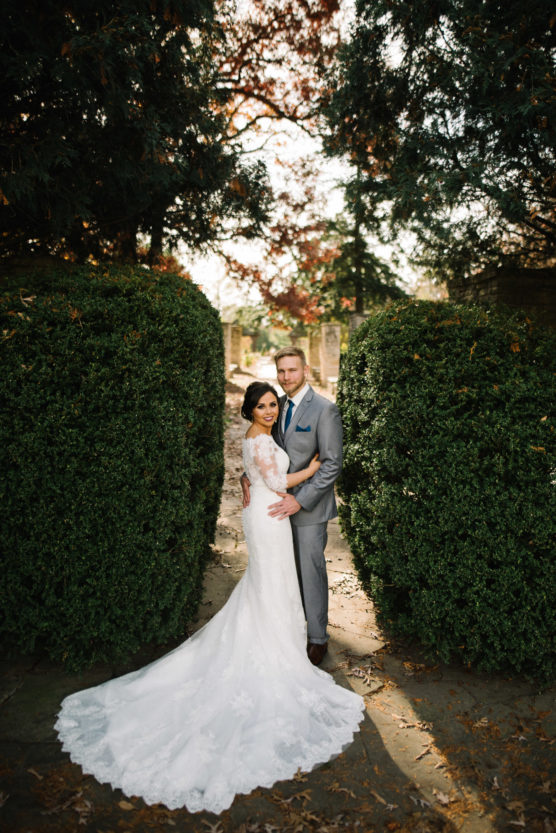 A bride in a white lace gown and a groom in a grey suit stand closely together on a stone path, surrounded by tall green hedges and trees, looking at the camera and smiling. by Justin Salem Meyer