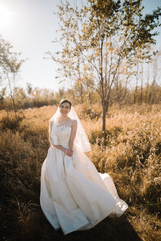 A bride in a white gown and veil stands smiling in a sunlit field with tall grass and trees, surrounded by warm, golden light on a clear day. by Justin Salem Meyer