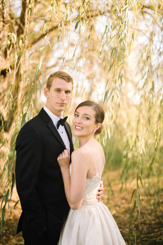 A young couple poses outdoors under willow branches; the man wears a black tuxedo and bow tie, while the woman in a white dress smiles, holding his lapel. Sunlight filters through the leaves, creating a warm, romantic atmosphere. by Justin Salem Meyer