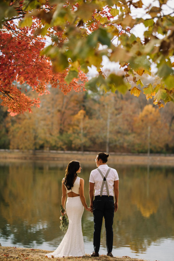 A bride and groom stand hand in hand by a serene lake, facing away, surrounded by colorful autumn trees. The bride wears a white dress and holds a bouquet, while the groom is in suspenders and a white shirt. by Justin Salem Meyer